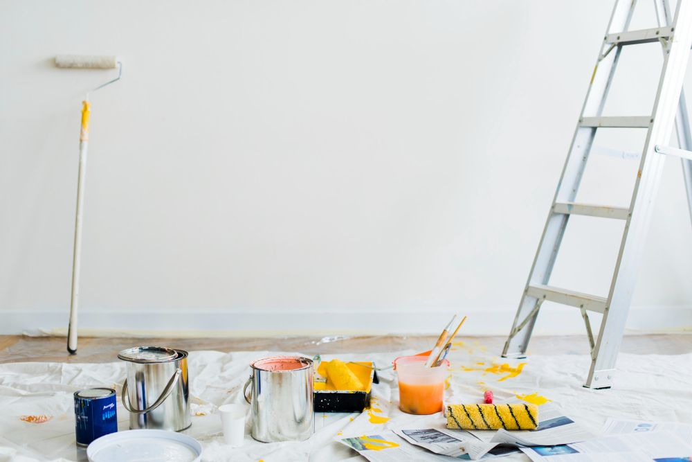 Painting supplies, paint cans, roller, and ladder set up against a white wall.