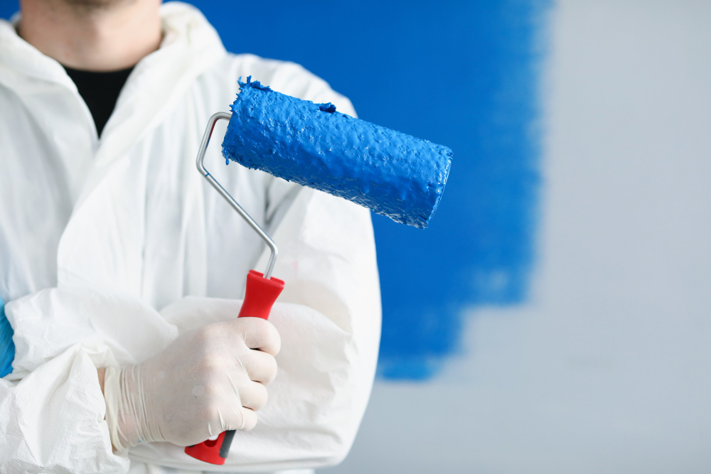 Person in white suit holds a blue paint roller against a partially painted blue wall.
