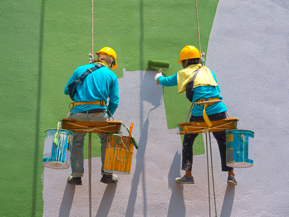 Two workers on harnesses paint a wall green while suspended by ropes.
