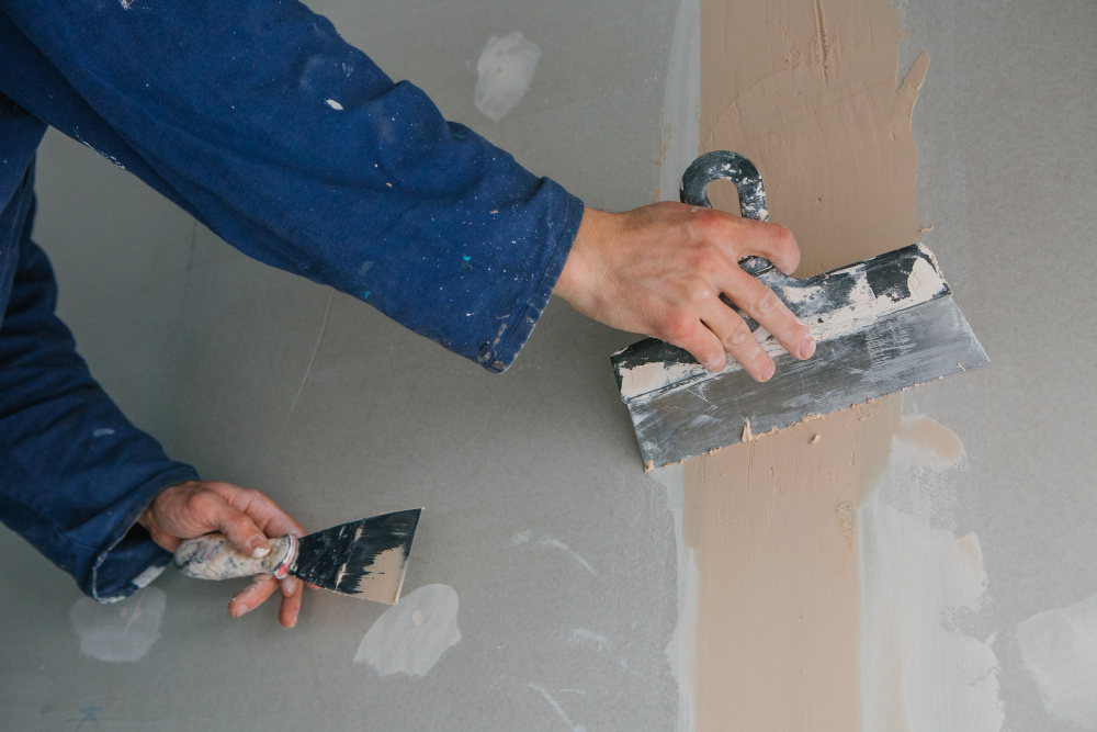 Person in blue using putty knives to spread plaster on a drywall seam.